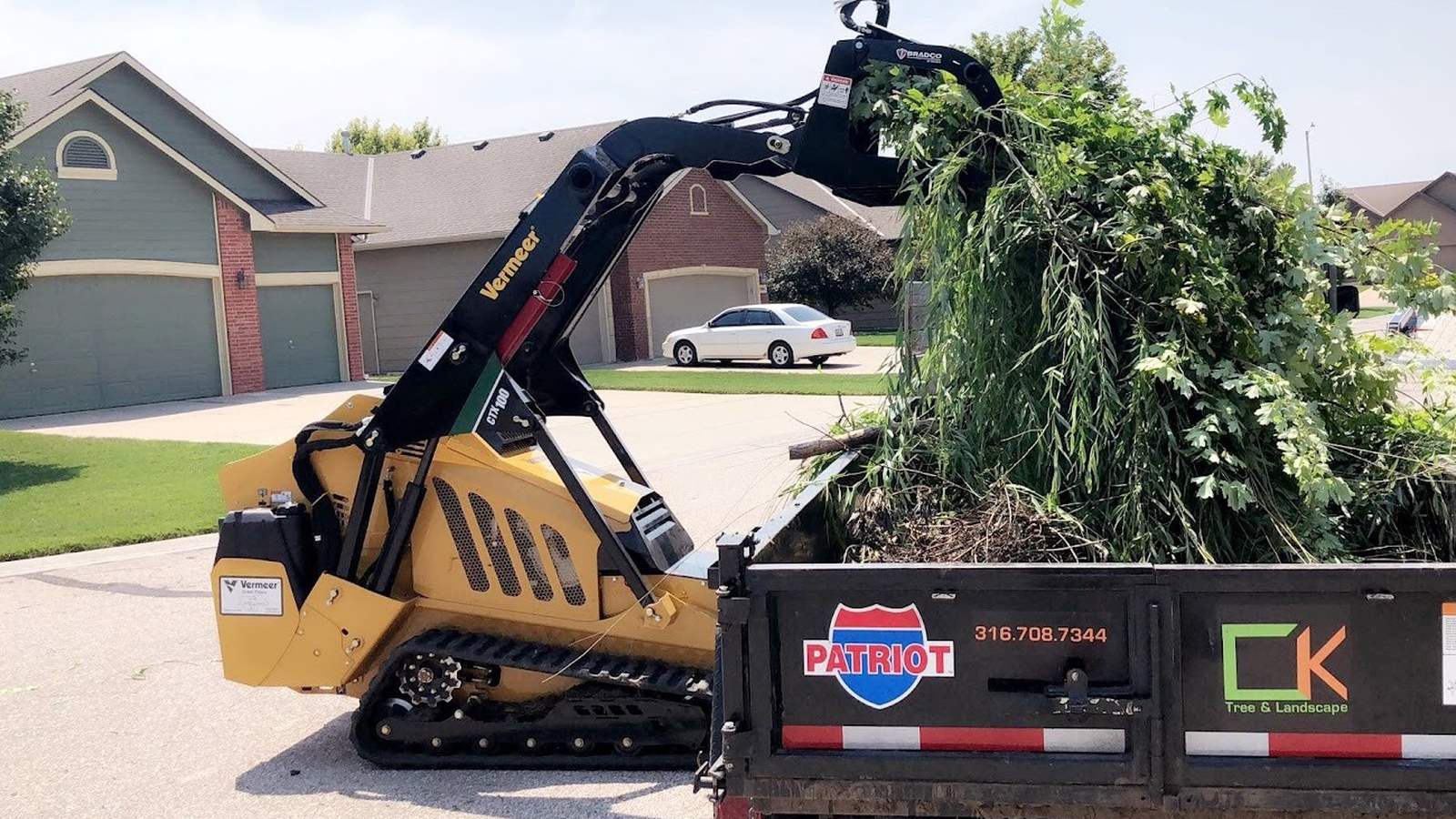A tracked mini loader lifting a tall pile of willow branches into a black dump trailer marked CK Tree & Landscape, on a suburban street.