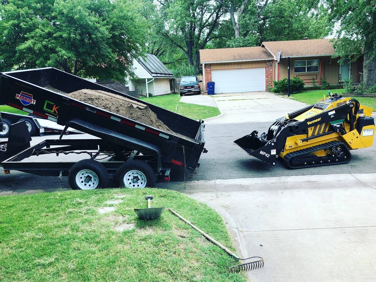 A black dump trailer tipped up with a heap of brown soil on a residential street, a yellow tracked mini loader and a rake and shovel on the grass nearby.